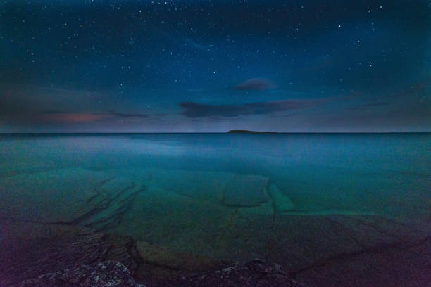 Milky way and starry sky with wispy clouds along the lakeshore of Georgian Bay at night with trees silhouetted and rocks in the clear water