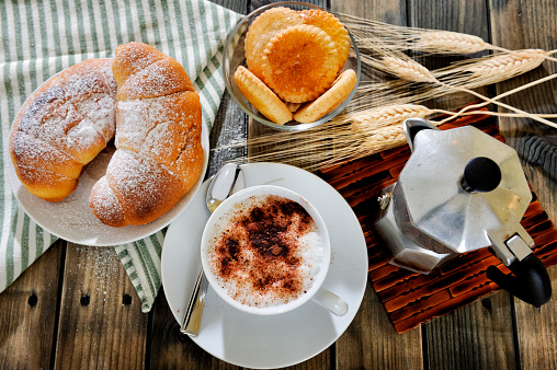 Top view of a cup of coffee with milk, croissants, cookies and the coffee pot on an antique wooden table.