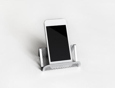 Cellphone with blank screen. Smartphone on metal stand at white paper background.