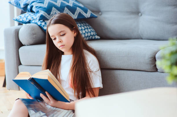 istock smart kid girl reading interesting book  at home, sitting with couch on background. Learning and education concept 930416936