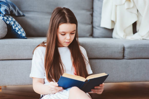 istock smart kid girl reading interesting book  at home, sitting with couch on background. Learning and education concept 930416948