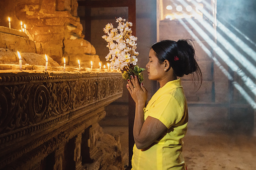 Burmese woman standing inside buddhist temple,praying to Buddha, holding a bouquet of flowers in her hands. Moody ambient natural light and light beams shining through the window of the temple pagoda.. Bagan, Mandalay, Myanmar, Asia.