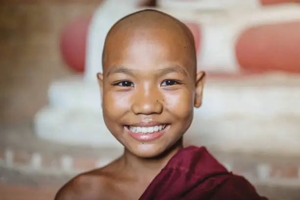 Happy burmese buddhist novice monk smiling towards the camera in front of huge buddha statue in stupa - temple in Old Bagan, Mandalay Region, Myanmar.