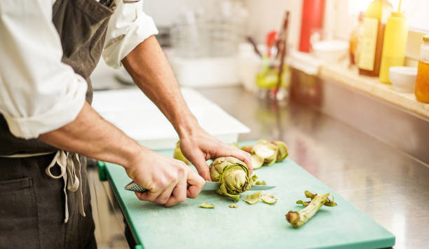 alcachofras de corte chef para a preparação do jantar - homem cozinhando dentro da cozinha do restaurante cozinha - foco na vegetal - vegan, estilo de vida e conceito de comida saudável - hotel commercial kitchen organic salad - fotografias e filmes do acervo