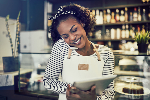 Smiling young African entrepreneur standing at the counter of her cafe talking on a cellphone and using a tablet