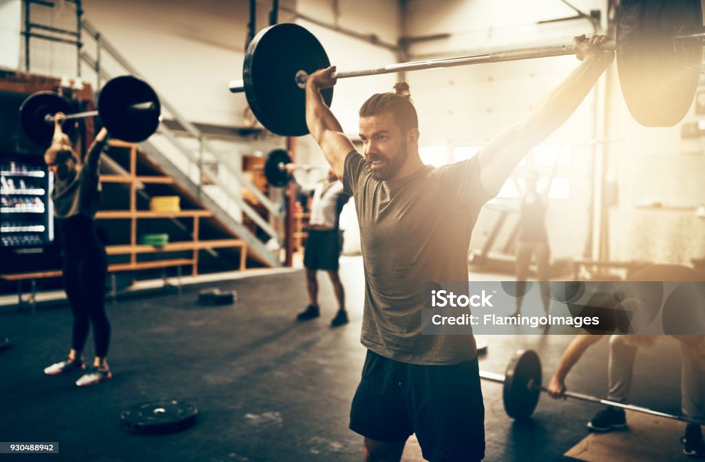 Fit young man weight training in a gym class Fit young man lifting heavy weights above his head during a workout class in a gym Weightlifting Stock Photo