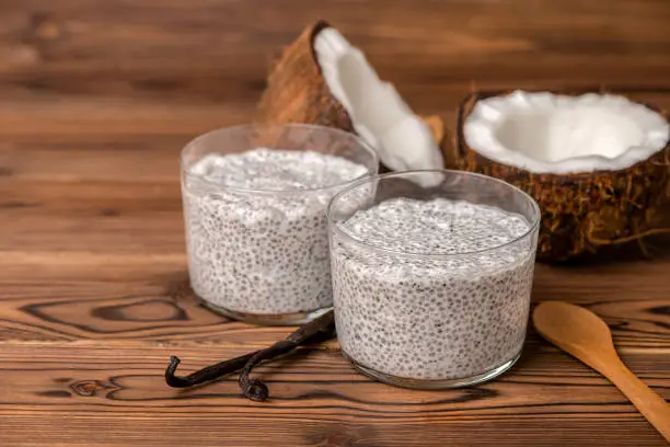 Photo of chia pudding in glass, decorated  vanilla pods, spoon and coconuts on wooden background, diet healthy eating and weight loss concept