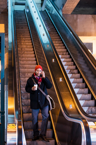 A mature African-American man dressed in warm clothing, going down an escalator, holding a disposable cup of coffee. He is a commuter on the way to work, leaving the train station, talking on his mobile phone.