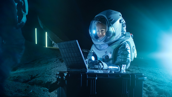 Mujer astronauta con traje espacial trabaja en un portátil, explorando nuevamente había descubierto planeta, comunicarse con la tierra. En el fondo su miembro de la tripulación y los Habitat del espacio. Concepto de colonización. photo