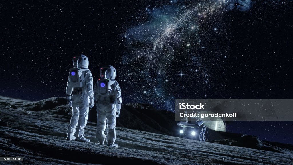 Two Astronauts in Space Suits Stand on the Planet and Looking at the The Milky Way Galaxy. In the Background Lunar Base with Geodesic Dome. Moon Colonization and Space Travel Concept. Moon Stock Photo