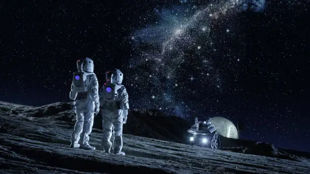 Photo of Two Astronauts in Space Suits Stand on the Planet and Looking at the The Milky Way Galaxy. In the Background Lunar Base with Geodesic Dome. Moon Colonization and Space Travel Concept.