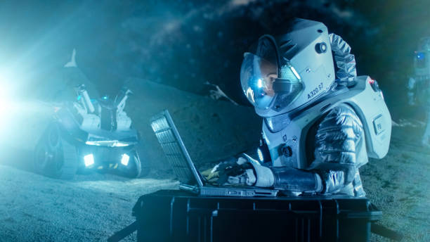 istock Female Astronaut Wearing Space Suit Works on a Laptop, Exploring Newly Discovered Planet, Communicating with the Earth. In the Background Her Crew Member and Space Habitat. Colonization Concept. 930523200