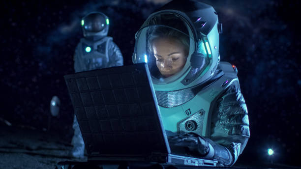 istock Female Astronaut Wearing Space Suit Works on a Laptop, Exploring Newly Discovered Planet, Communicating with the Earth. In the Background Her Crew Member and Living Station. Extraterrestrial Colonization Concept. 930523250