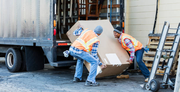Two workers with a truck, moving large box Two multi-ethnic mature workers in their 40s at the back of a truck, loading or unloading a large cardboard box. The men are wearing plaid shirts, reflective vests and jeans. They are moving merchandise for a furniture store. unloading photos stock pictures, royalty-free photos & images