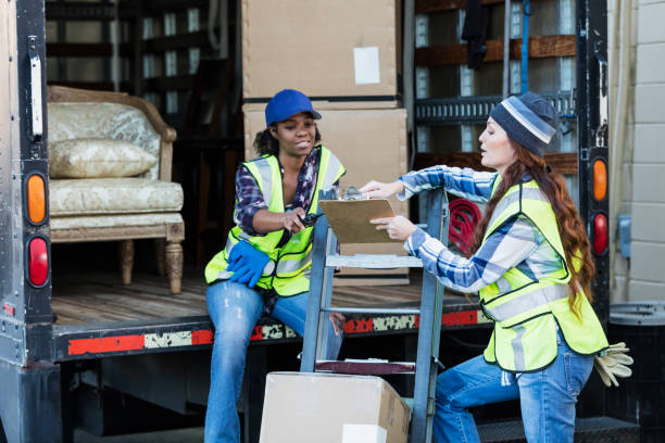 Two female workers with a truck, moving furniture, boxes Two multi-ethnic female workers in their 30s and 40s at the back of a truck, one holding a clipboard. The women are wearing reflective vests and jeans. They are moving merchandise for a furniture store. mover delivery person box hand truck stock pictures, royalty-free photos & images
