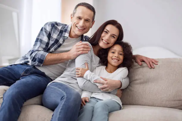 Happy together. Beautiful content curly-haired girl smiling and sitting on the couch with her parents and they hugging each other