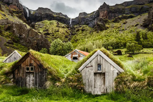 Photo of Icelandic turf houses and rocky canyon with waterfall in the background near Kalfafell vilage, South Iceland