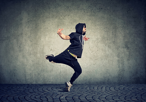 Stylish young man in trendy sportive clothing performing street dance with on gray wall background.