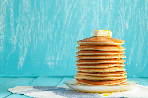 Stack of pancakes with honey and piece of butter on plate which stands on napkin on blue wooden table. Copyspace. Concept of shrovetide treats. Selective focus