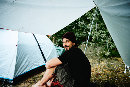 The male adult is sitting under a tarp and next to his tent at a campsite at Lake Vänern in Sweden. He is smiling and looks into the camera while waiting for the rain to stop.