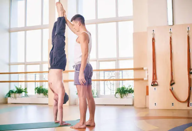 Photo of Yoga practice, instructor helping student to do handstand in class, Adho Mukha Vrikshasana, Downward facing Tree Pose