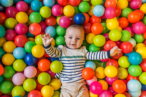 Happy Caucasian laughing baby boy having fun in ball pit on birthday party in kids amusement park and indoor play center and looking at camera. Photo of Child playing with colorful balls in playground ball pool during the day. Activity toys for little kid.