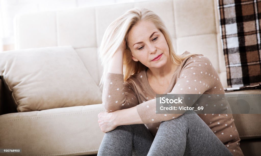 Femme aux pieds nus, assis à l’étage embrasser ses genoux, près de canapé à la maison, sa tête vers le bas, s’ennuie, troublée par la violence domestique d’âge moyen. - Photo de Femmes d'âge mûr libre de droits