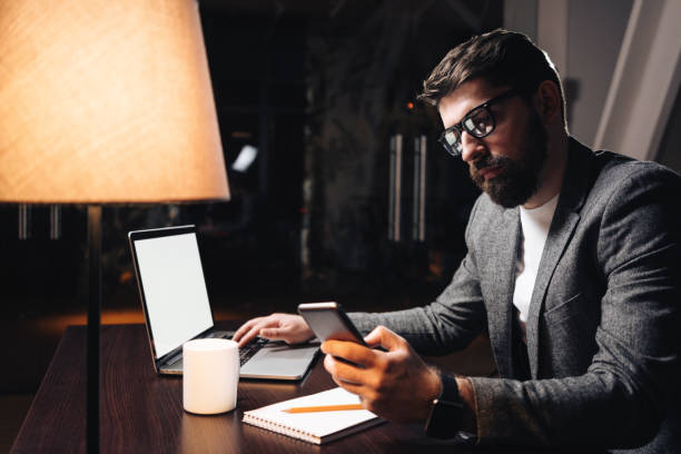 istock Businessman using smartphone and laptop in coworking studio. Young bearded man working about new startup at night in modern loft office. Film effect 930788530
