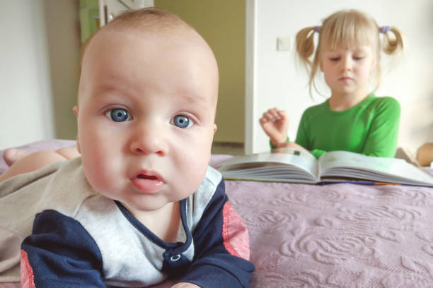 ritratto di bambino pensieroso che fa selfie su un letto. sorella che legge il libro sullo sfondo.  concetto di diversi interessi di età - pensive only baby girls baby girls baby foto e immagini stock