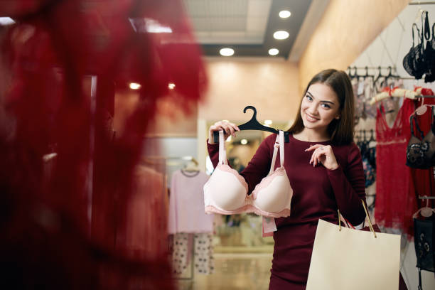 Young woman chooses sexy brassiere among set in a boutique. Pretty girl considers bra with shopping bags in her hand in underwear store stock photo