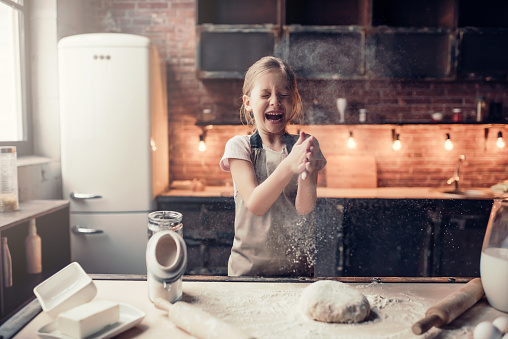 Little cute girl on kitchen is ready for cooking. Laughing while clapping hands with flour.