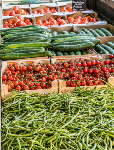 A vibrant vegetable market stall in Palermo, Sicily, featuring fresh green beans, red tomatoes, and cucumbers. A hand packaging vegetables adds a human touch to this scene of local market life.