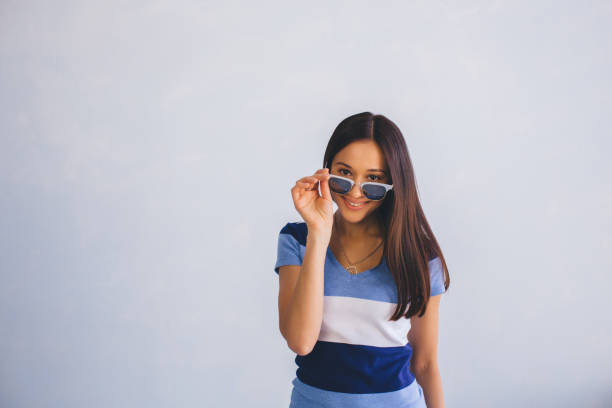 latino woman wearing casual clothes looking through sunglasses and smiling, while standing against background with copy space. Positive youth