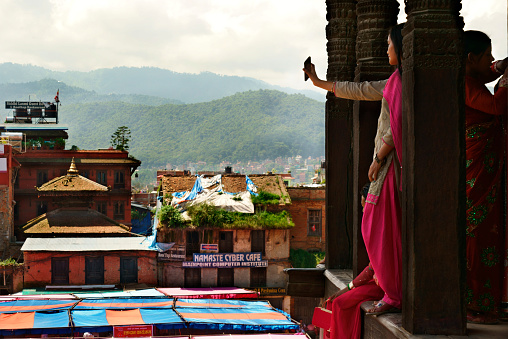 Bhaktapur, Nepal - August 24, 2017: Young nepalese woman clad in traditional clothing standing on top of Nyatapola temple looking at the panoramic view of the city and the surrounding mountains and taking photographs using her mobile phone. The women wearing colorful clothing typical for the Kathmandu valley and the Hindu culture especially on religious events like the Teeji festival in August. Nyatapola temple at Taumadhi square in old town Bhaktapur. Hinduism. Asian culture, spirituality and religion. XXXL (Sony Alpha 7R)