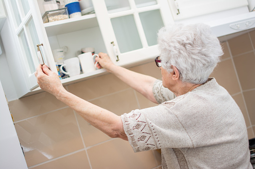 Back view of senior woman taking dishes from cupboard in the kitchen.