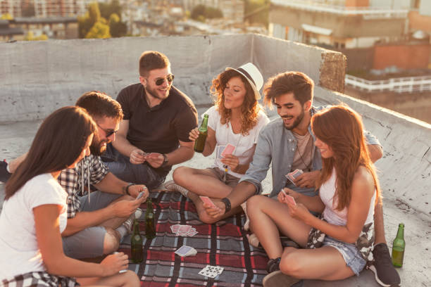 Friends playing cards Group of young people sitting on a picnic blanket, having fun while playing cards on the rooftop. Focus on the girl in the middle friends playing cards stock pictures, royalty-free photos & images