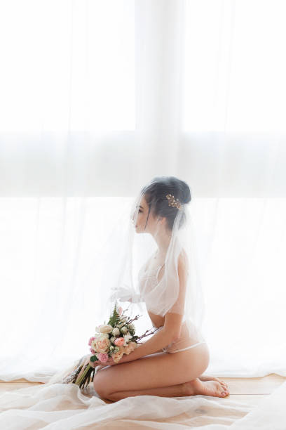 young bride in a white lingerie sitting on the floor next to the big window, covered with veil, morning preparation before wedding - sensuality sex symbol dressing room women imagens e fotografias de stock