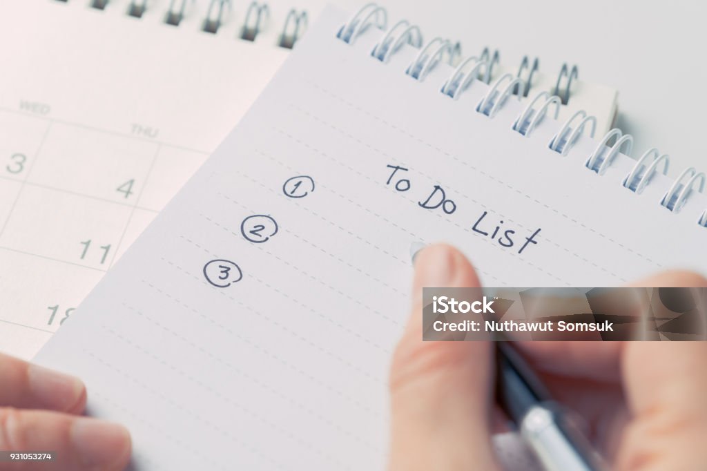 Weibliche Hand mit schwarzer Feder schreiben To Do List von Zahl auf weißem Papier Notizblock mit Desktop-Kalender-Datum auf weißer Bürotisch priorisiert - Lizenzfrei Aufgabenliste Stock-Foto