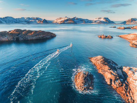 Aerial view of fishing boats, rocks in the blue sea, snowy mountains and colorful sky with clouds at sunset in winter in Lofoten islands, Norway, Landscape with two ship, blue water, waves. Top view