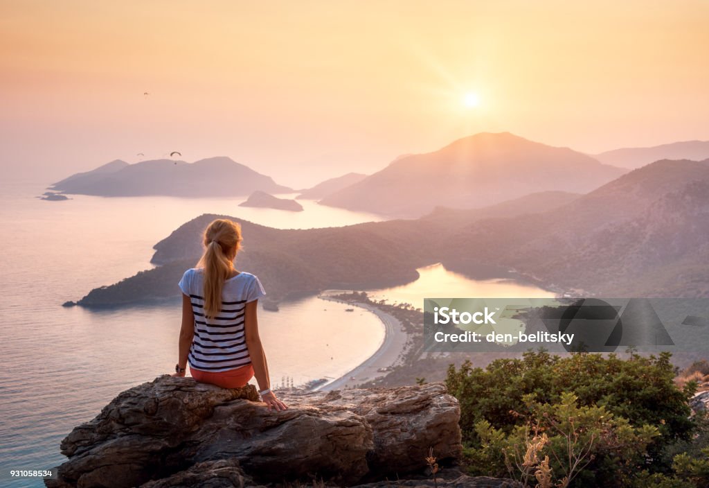 Landscape with girl, sea, mountains and orange sky Young woman sitting on the top of rock and looking at the seashore and mountains at colorful sunset in summer. Landscape with girl, sea, mountain ridges and orange sky with sun. Oludeniz, Turkey. Beach Stock Photo