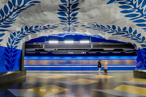 Stockholm, Sweden - August 28, 2017: People standing in front of a moving train on a platform of the underground metro or tunnelbana central station T-Centralen with updated intricate wall  designs from 1975