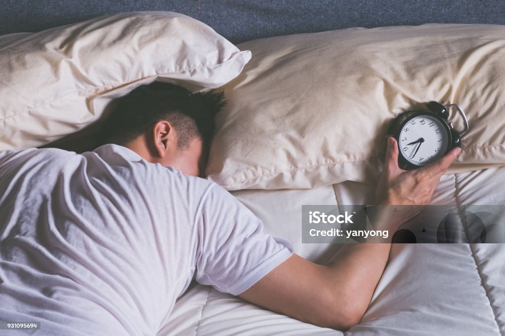 sleeping asian young male disturbed by alarm clock early morning in bed Waking up Stock Photo