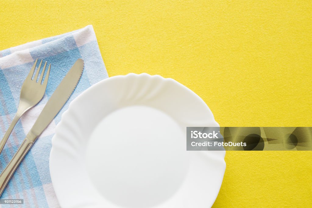 Clean, empty, white plate, fork and knife with yellow tablecloth and blue towel on a table. Cutlery concept. Flat lay. Top view. Free space for text or a logo. Plate Stock Photo