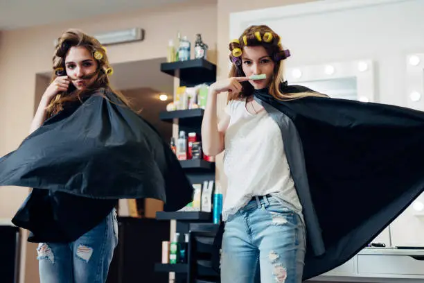 Photo of Two female friends goofing around in front of the camera making moustache from hair and curler standing in flying cape like heroes at beauty shop