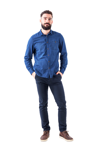 Relaxed young man wearing blue denim shirt with hands in pockets looking at camera. Full body isolated on white background.