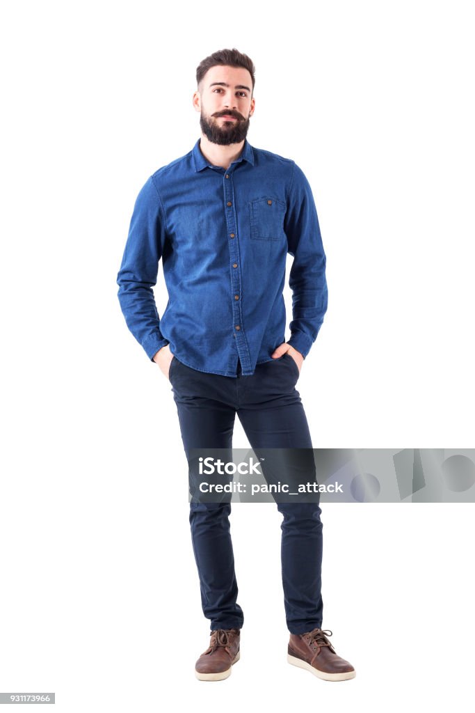 Relaxed young man wearing blue denim shirt with hands in pockets looking at camera Relaxed young man wearing blue denim shirt with hands in pockets looking at camera. Full body isolated on white background. Men Stock Photo