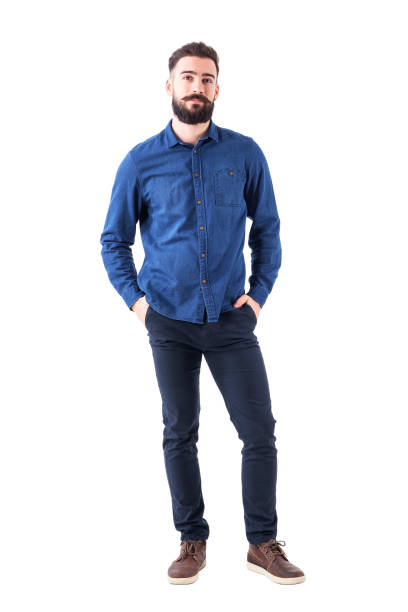 Relaxed young man wearing blue denim shirt with hands in pockets looking at camera Relaxed young man wearing blue denim shirt with hands in pockets looking at camera. Full body isolated on white background. business casual fashion stock pictures, royalty-free photos & images