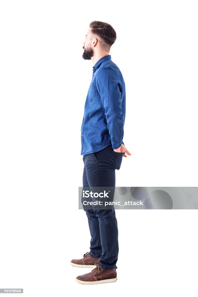 Vista laterale di giovane uomo barbuto elegante con le mani in tasca dietro in piedi e guardando - Foto stock royalty-free di Persone