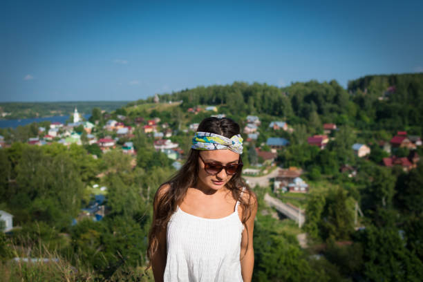 Young woman in sunglass and bandage on the head stands on a hill in warm summer day and looking down. Beautiful girl is standing in the park on the background of a small village and green trees