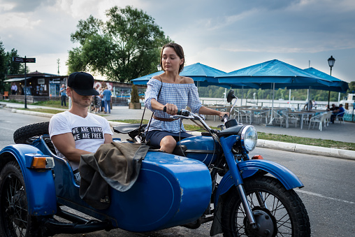 Young couple is traveling on a retro motorcycle. Beautiful woman with boyfriend went in road trip. Serious girl is driving motorcycle. We are young we are free. Man is relaxing in the sidecar